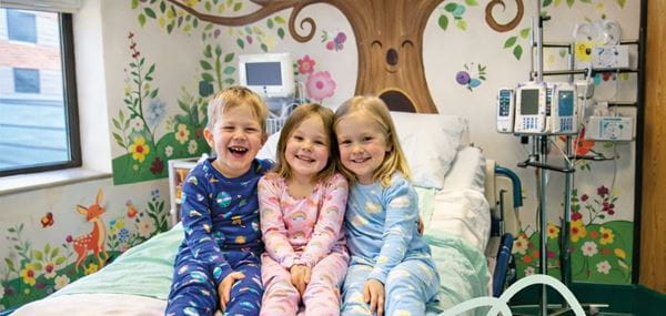 Three children sit on a hospital bed in a nature-themed room.