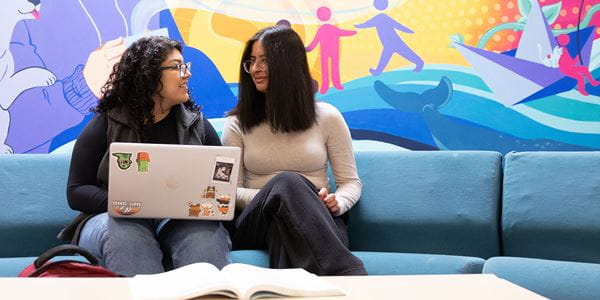 Two students sitting on couch in front of mural.